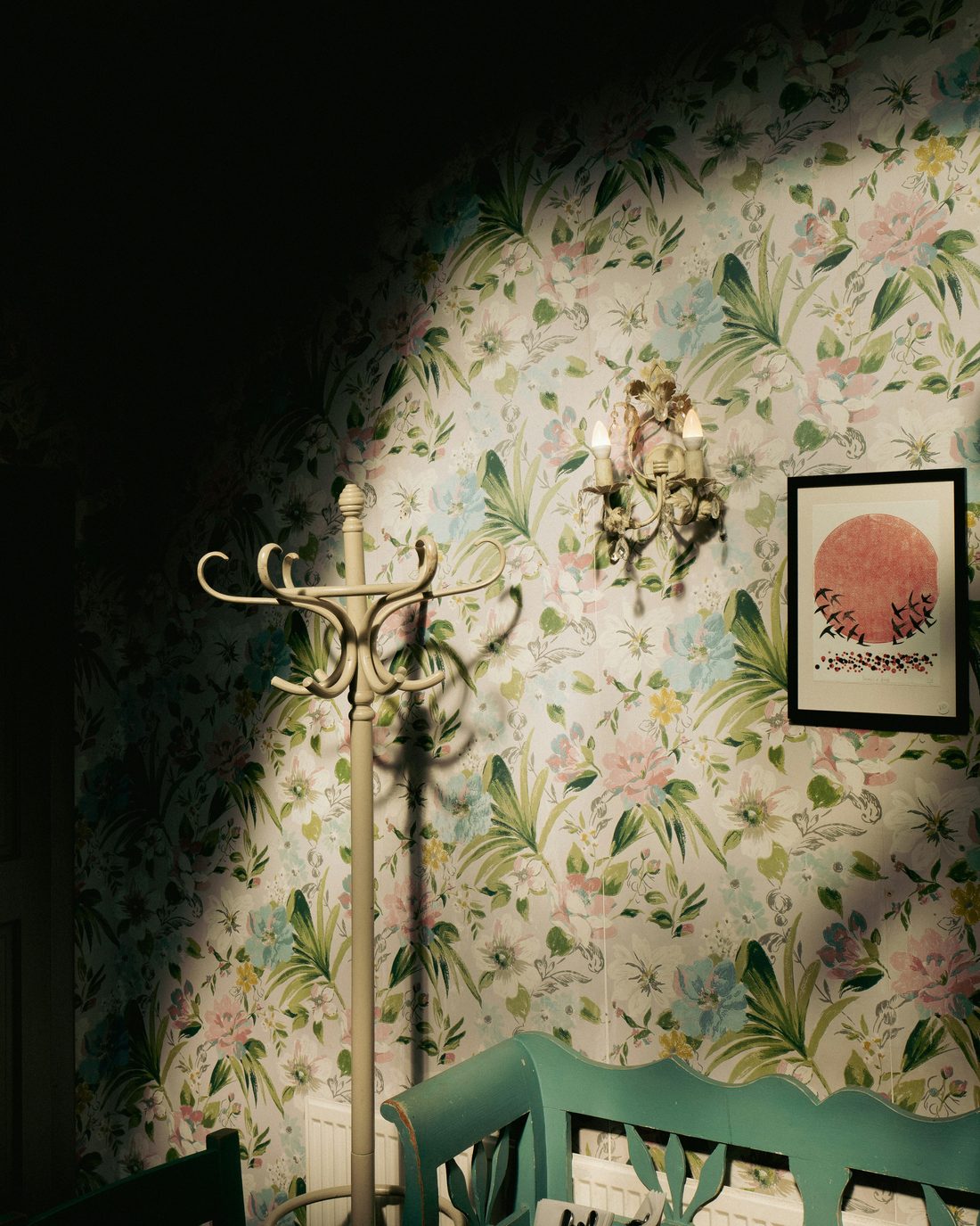Pale botanical wallpaper with pink blooms and green palms in a softly lit entry with a bentwood coat stand, wall sconce and teal bench