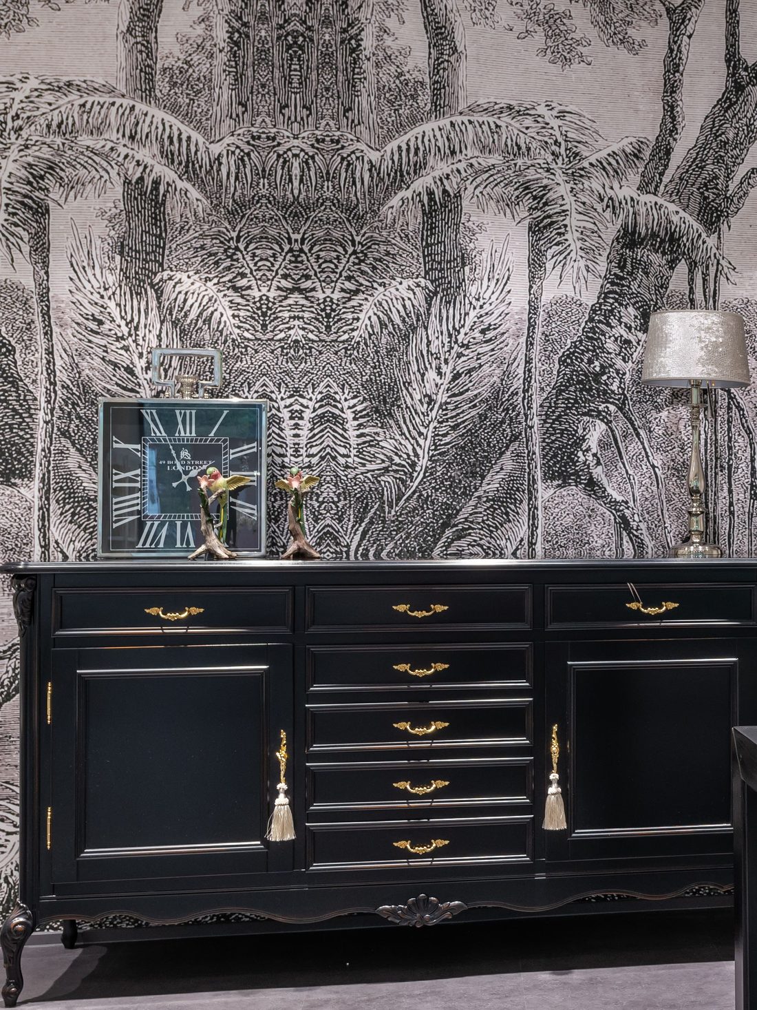 Black and white engraved-style mural of palms and trees behind a black lacquered sideboard with brass handles, a clock and a silver lamp
