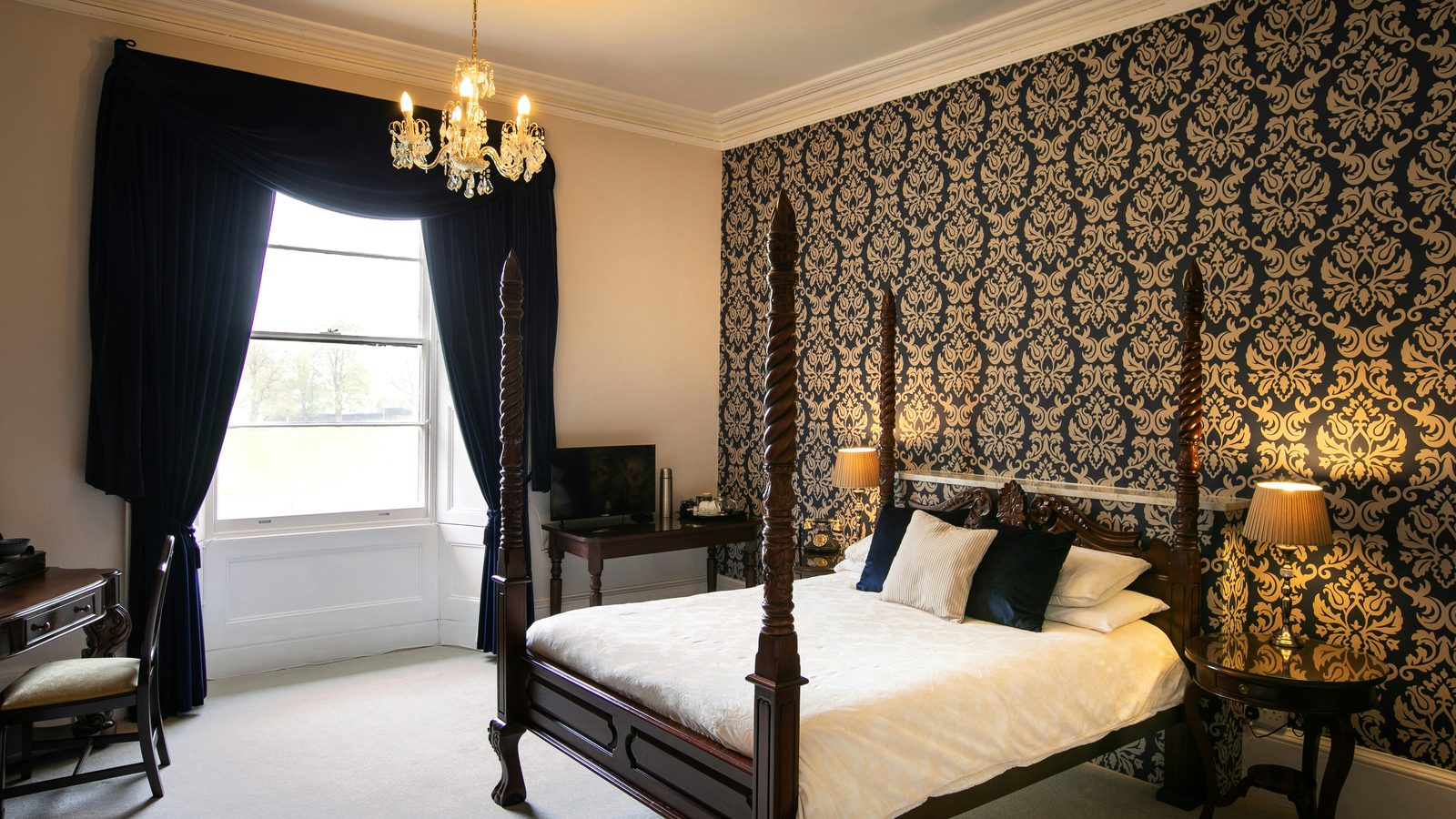Elegant bedroom with a navy and gold damask wallpaper feature wall, a carved four-poster bed, crystal chandelier and dark velvet drapes