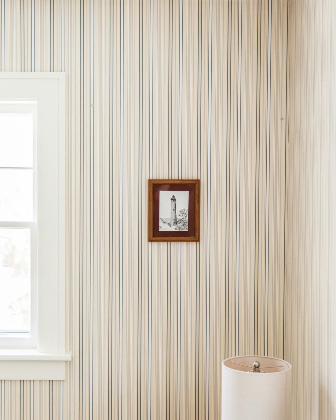 Calm room with fine cream, blue and grey striped wallpaper, a small framed lighthouse print and a white window frame