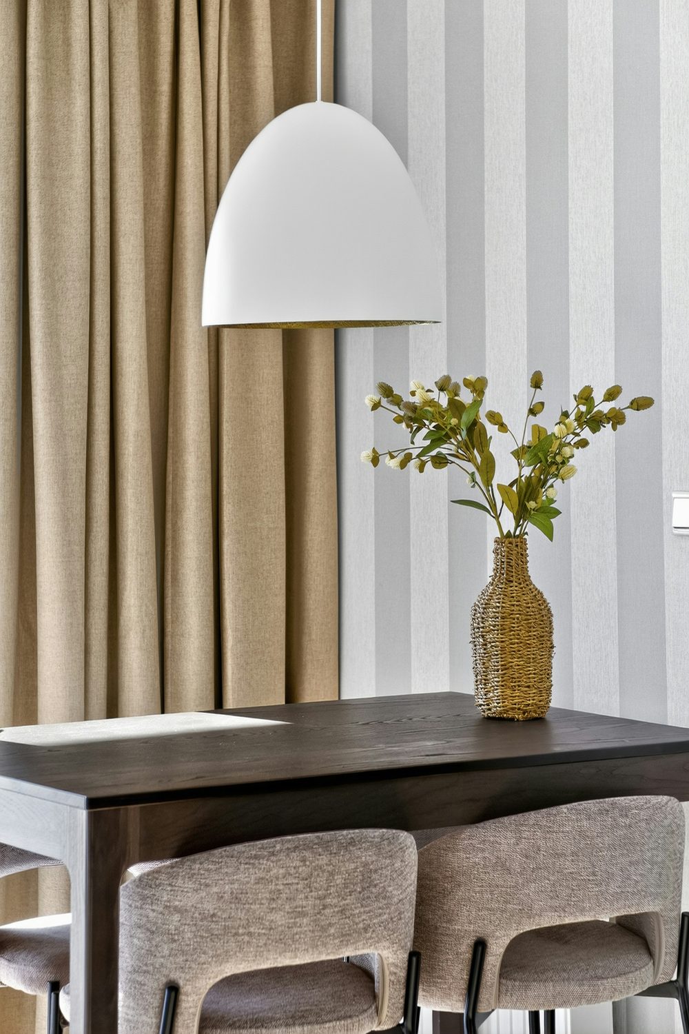 Contemporary dining corner with grey and white striped wallpaper, a white pendant lamp, a dark table and a vase of greenery