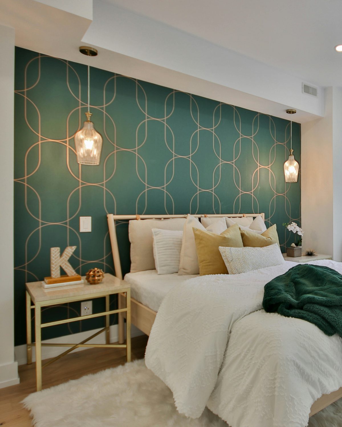 Bedroom feature wall in deep green wallpaper with a fine gold geometric arch pattern behind a bed with gold and emerald cushions