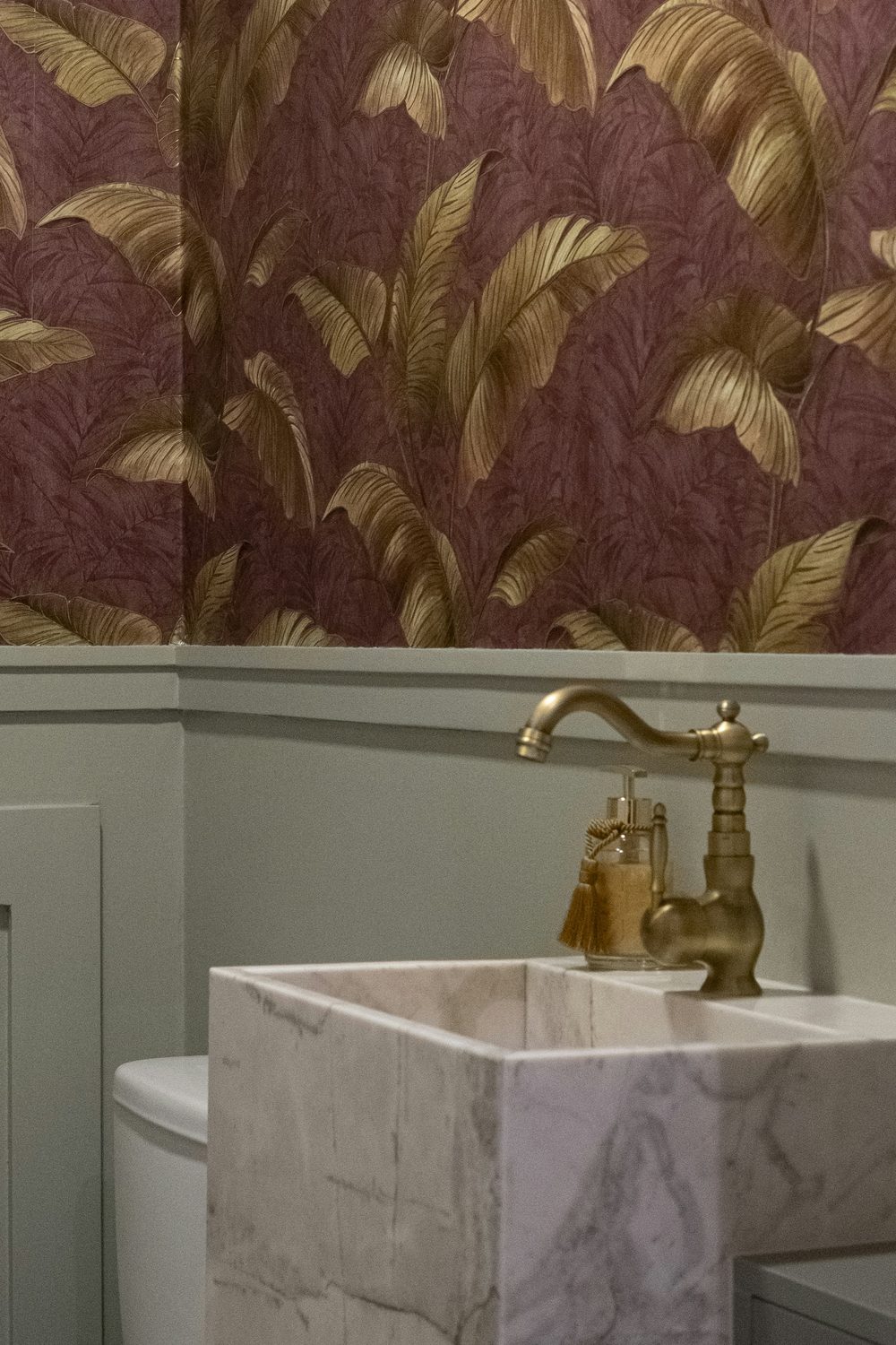Powder room with plum wallpaper patterned in large metallic gold leaves above sage-green panelling, a brass tap and a marble basin